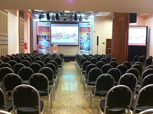 Salão vazio montado em auditório, com cadeiras em fileiras, palco, telões e iluminação