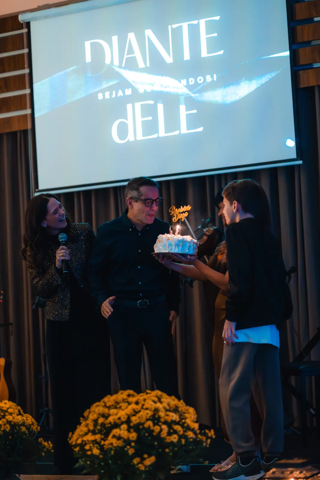 Comemoração no palco com bolo e velas acesas, diante do telão e de arranjos de flores