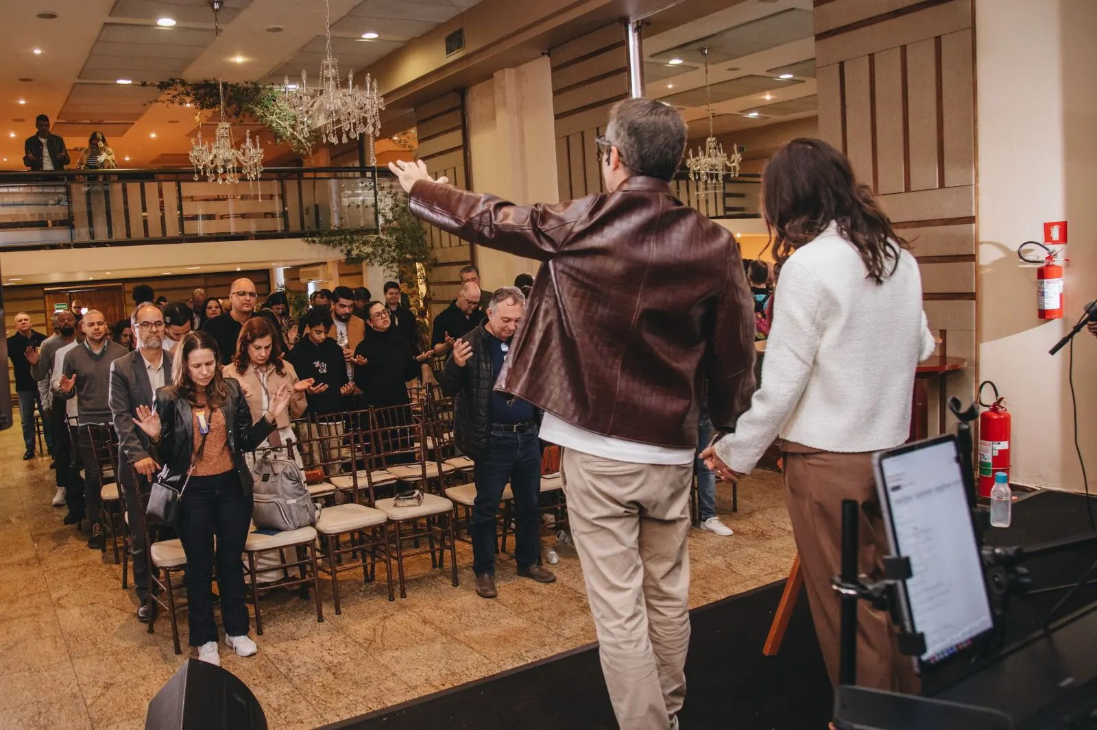 Convidados de pé no salão diante do palco, com lustres e mezanino ao fundo