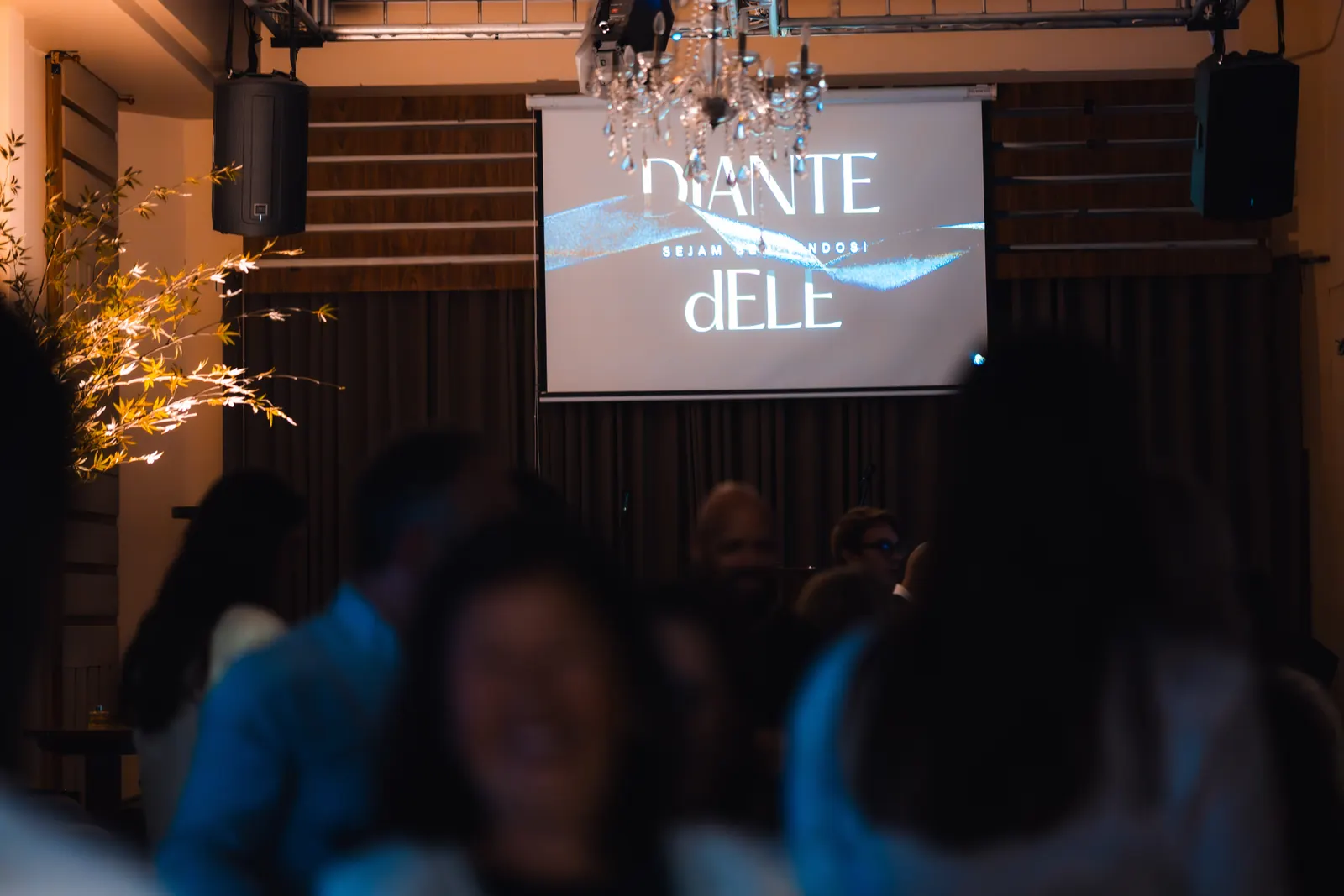 Convidados no salão da Marvelous Casa diante do palco, com telão e lustre ao fundo